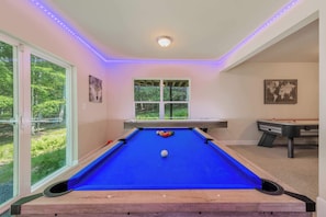 Een pooltafel met een blauwe viltkleed, omringd door muren met een raam en een deur, en een plafond met LED-verlichting.