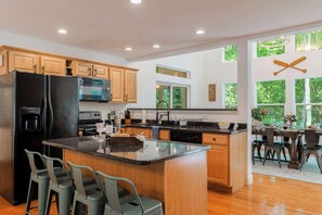 A modern kitchen with a central island, wooden cabinets, a black refrigerator, and a dining area with a view of trees.