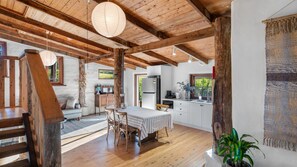 A rustic interior with exposed wooden beams, a dining area with a table and chairs, a kitchen with white cabinets, and a large window. - The Cottage at Indooroopilly (Bellawongarah)