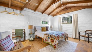 Ein Schlafzimmer mit Holzbalkendecke, einem Bett mit floralem Bettzeug, einem Holzstuhl, einem kleinen Tisch mit einer Lampe und einem Fenster mit Vorhängen.