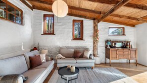 A cozy living room with a sofa, a coffee table, and a wooden cabinet. There are two windows with wooden frames and a hanging light fixture. - The Cottage at Indooroopilly (Bellawongarah)