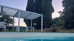 En swimmingpool med et overdækket opholdsområde og høje træer i baggrunden.