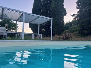 Ein Pool mit überdachter Liegefläche und hohen Bäumen im Hintergrund.