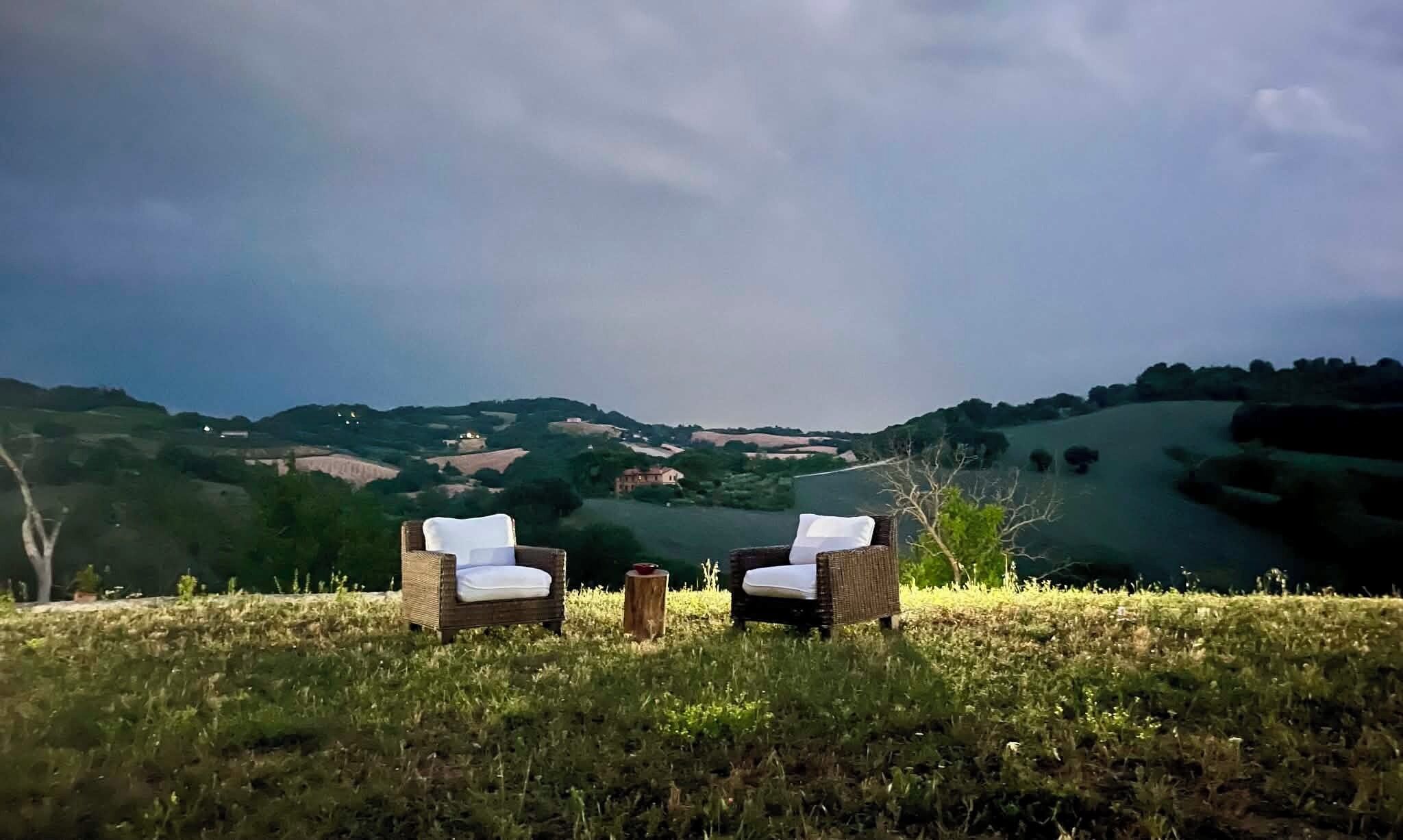 Two wicker chairs with white cushions on a grassy field, overlooking rolling hills and a distant building.