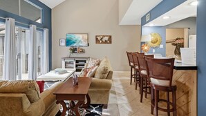 Uma sala de estar moderna com sofá, mesa de centro e área de jantar com cadeiras. Há uma TV fixada na parede e uma peça decorativa.