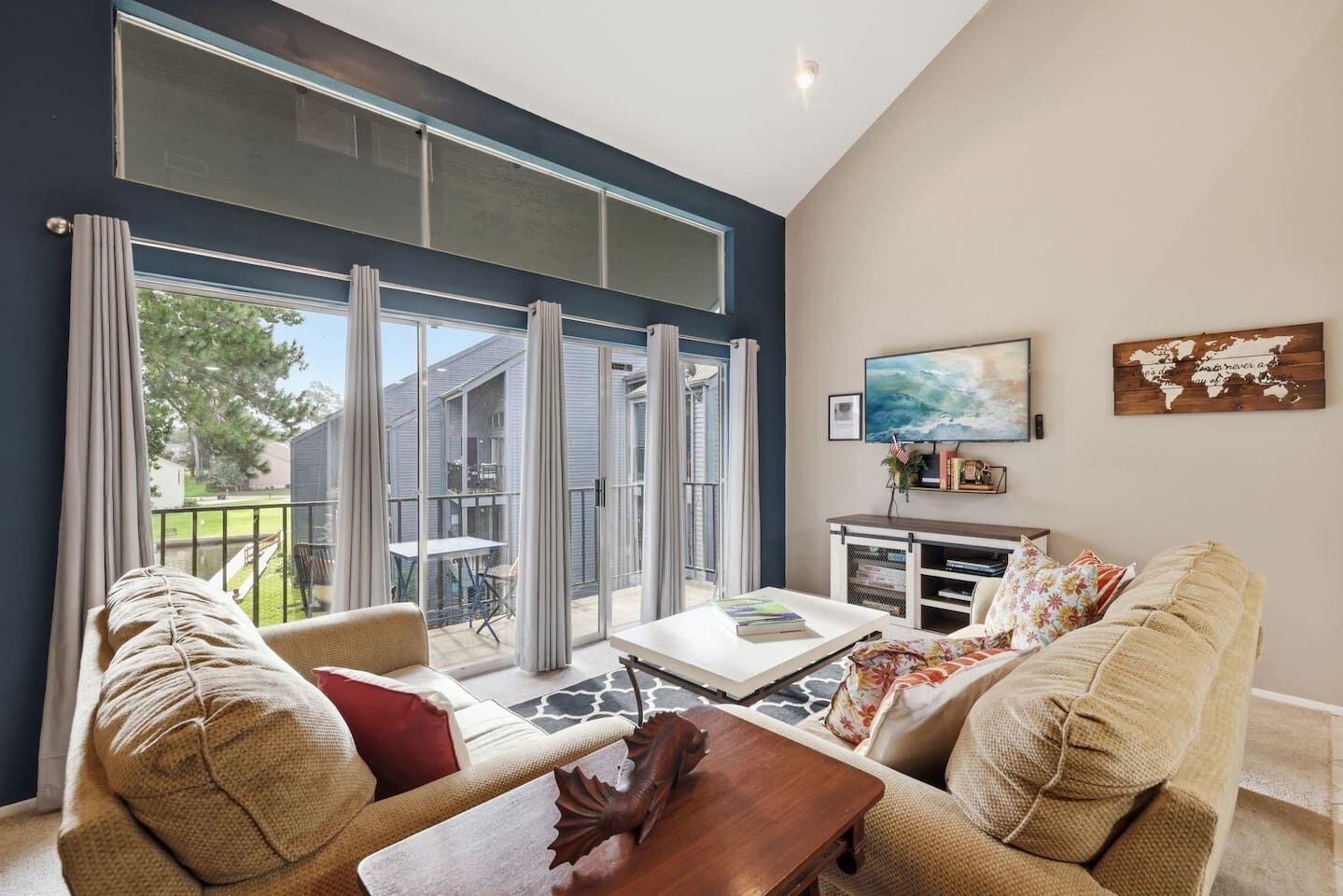 A spacious living area with a large sliding glass door leading to a balcony, a comfortable sofa set, a wooden coffee table, and a flat-screen TV mounted on the wall.