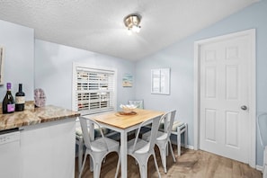A kitchen with white cabinets, a wooden dining table, and white chairs. There is a window with blinds, a door, and a ceiling light fixture. - 1002 Seaside Cottage (Myrtle Beach)