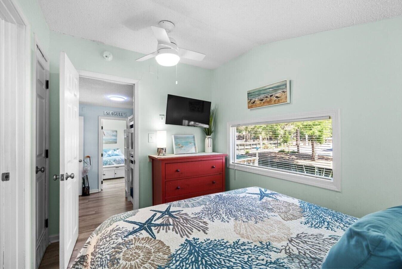 A bedroom with a bed, a red dresser, a ceiling fan, a TV, and a window with a view.