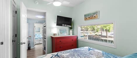 A bedroom with a bed, a red dresser, a ceiling fan, a TV, and a window with a view.