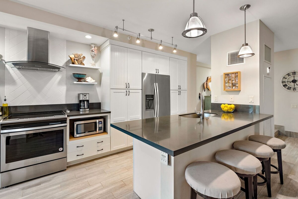 A modern kitchen with stainless steel appliances, a central island with bar stools, pendant lighting, and a clock on the wall.