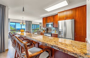 A modern kitchen with wooden cabinets, stainless steel appliances, a granite countertop, and a view of the ocean. - The Kaanapali Alii by Maui Resort Rentals (Kaanapali)