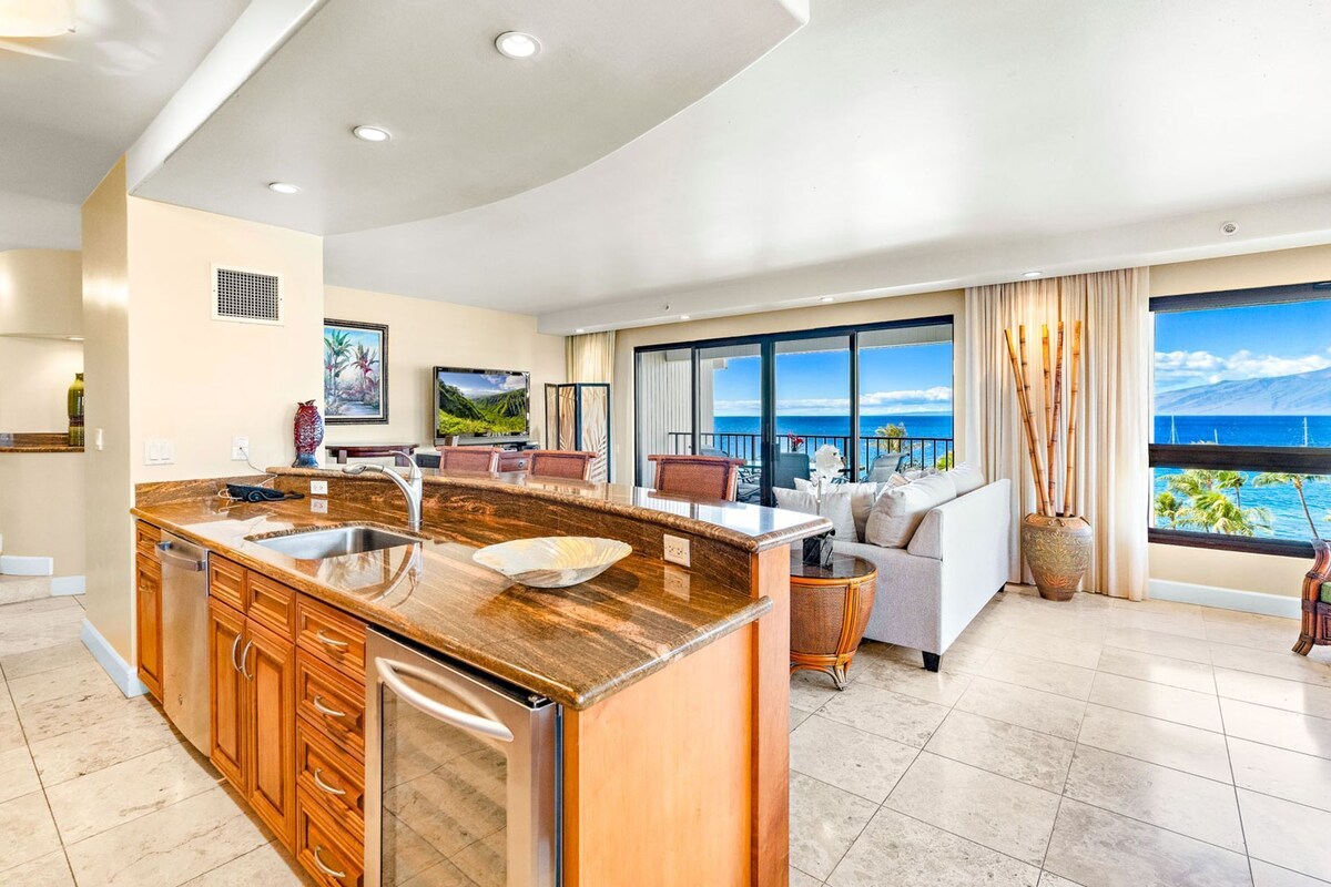 A modern kitchen with a central island, a dining area with a sofa and chairs, and a large balcony with a view of the ocean.