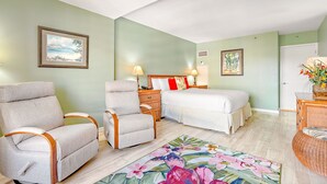 A bedroom with a bed, two armchairs, a nightstand, a lamp, a floral rug, and a door. - The Kaanapali Alii by Maui Resort Rentals (Kaanapali)