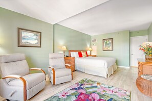 A bedroom with a bed, two armchairs, a nightstand, a lamp, a floral rug, and a door. - The Kaanapali Alii by Maui Resort Rentals (Kaanapali)
