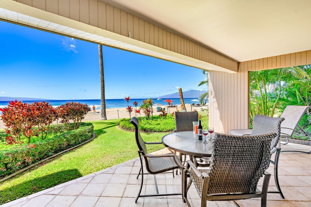 A covered patio with a table and chairs, overlooking a beach with palm trees and a clear blue sky.