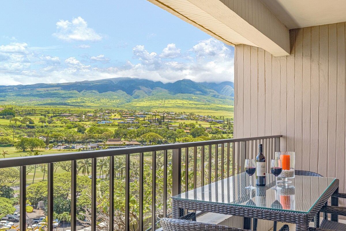 A balcony with a glass table, a bottle of wine, and glasses, overlooking a green landscape with houses and mountains.