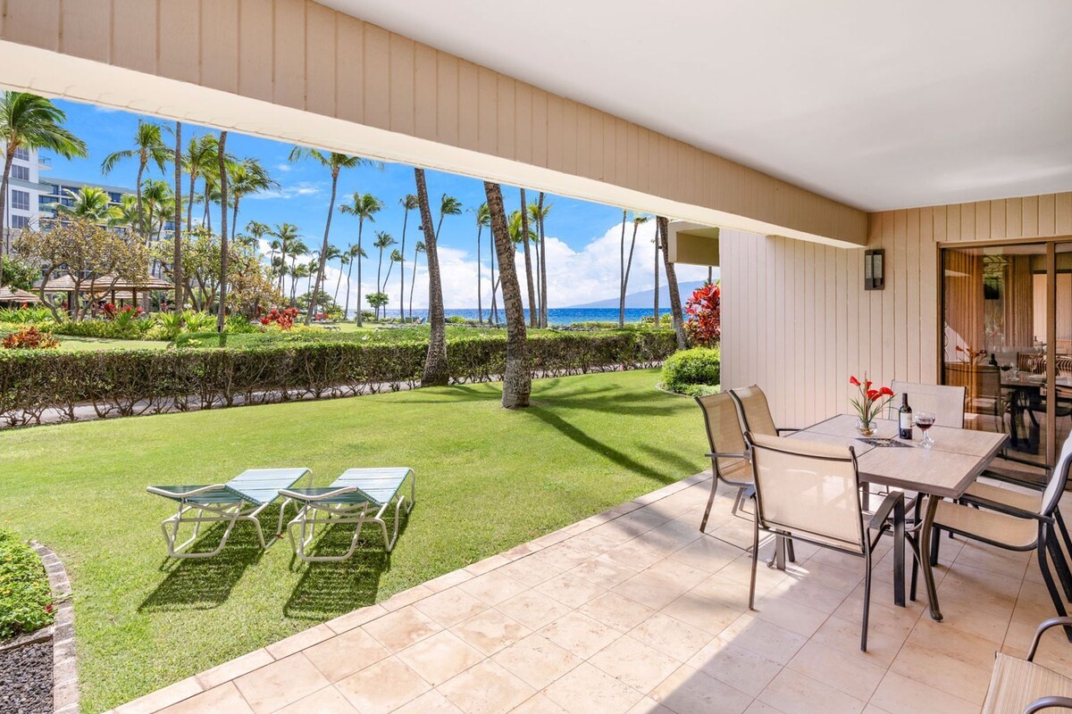 A patio with a table and chairs, a lawn, and a view of palm trees and a building across the water.
