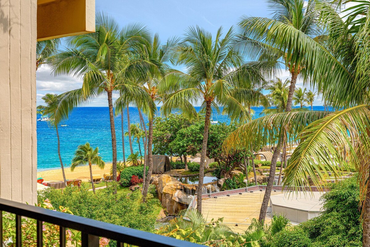 A balcony with a view of a beach, palm trees, and a pool area.