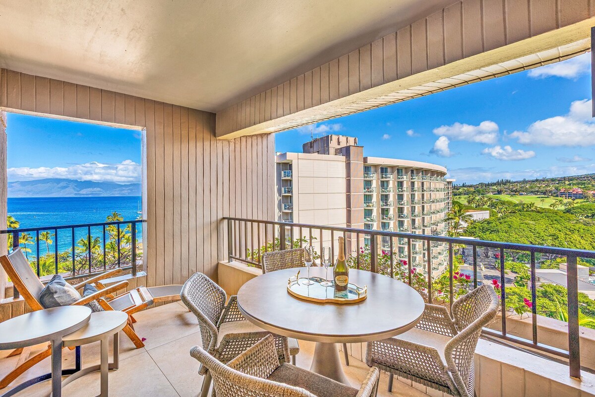 A balcony with wicker furniture, a round table, and a view of a hotel and ocean.