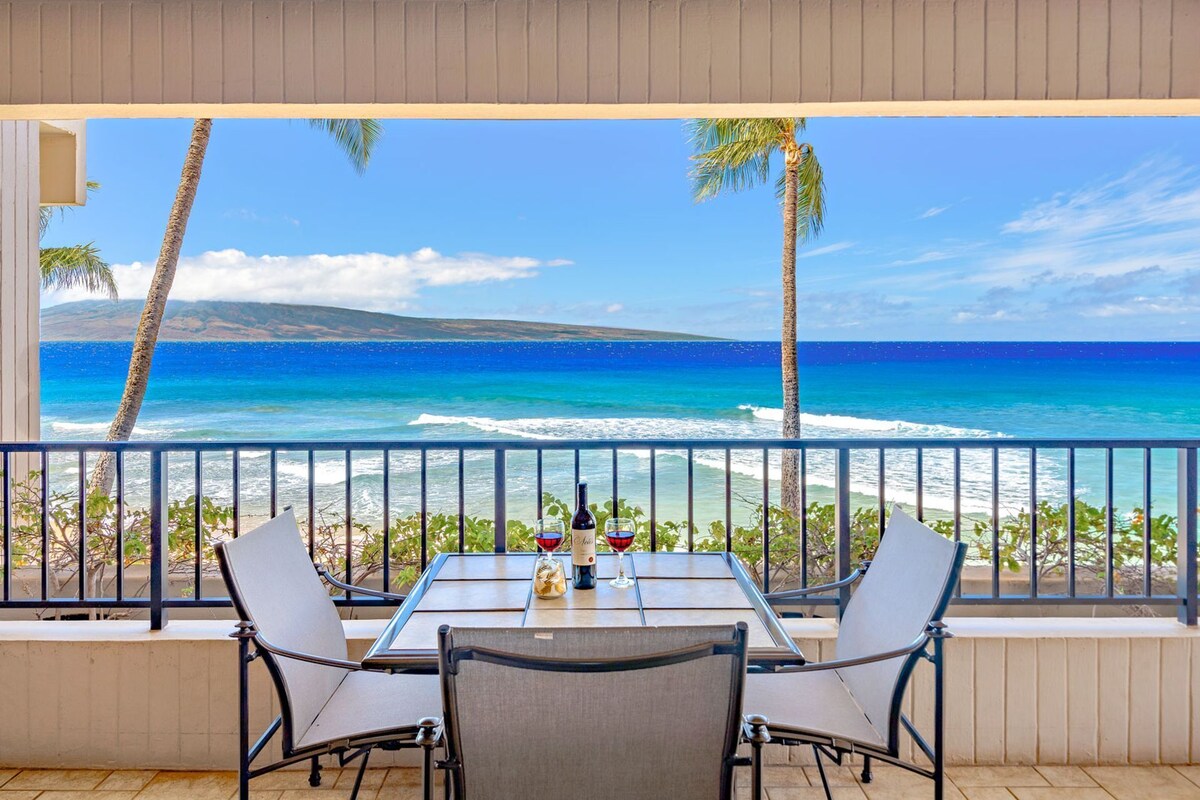 A balcony with a table and chairs overlooking a beach with palm trees and the ocean.