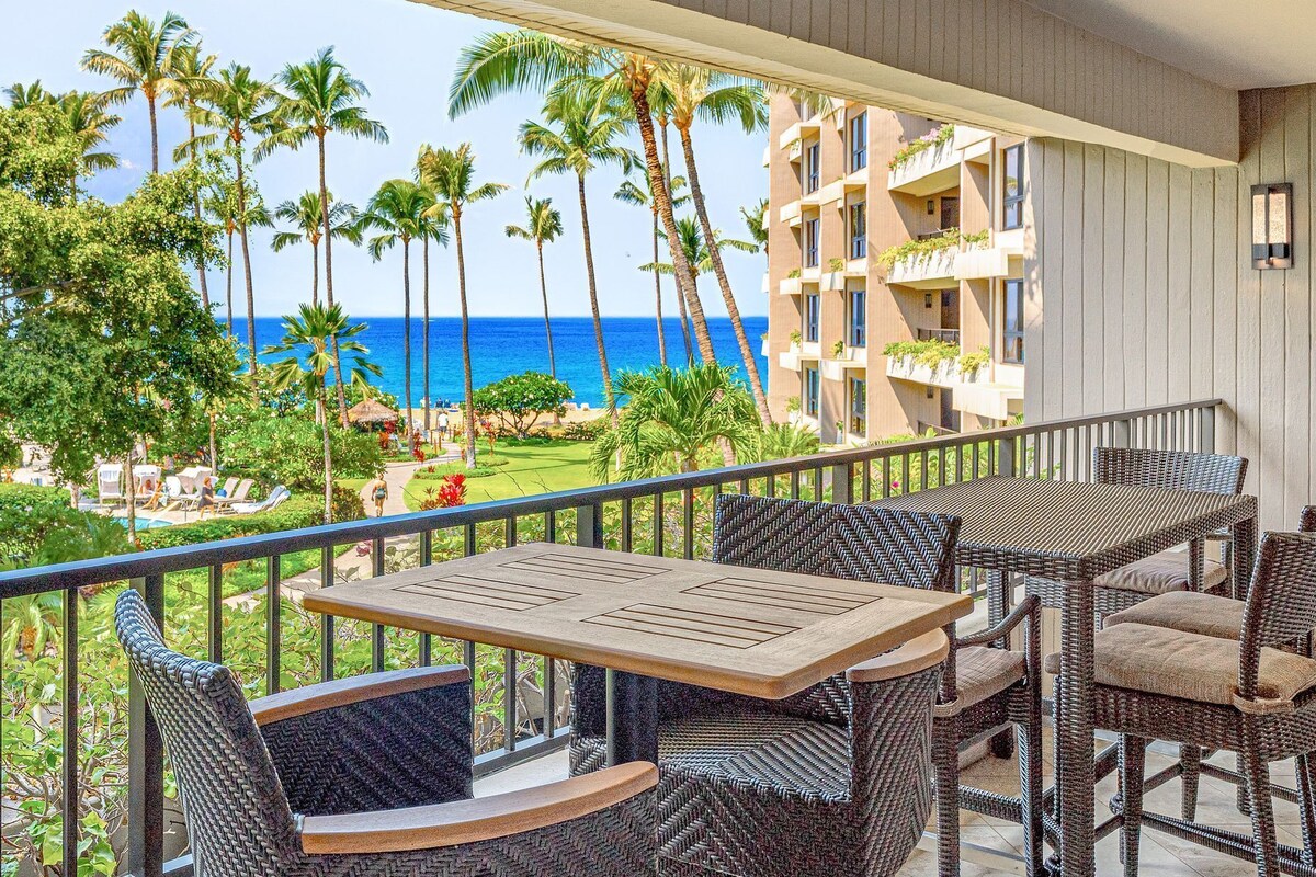 A balcony with wicker furniture overlooking a beach with palm trees and a building with balconies.