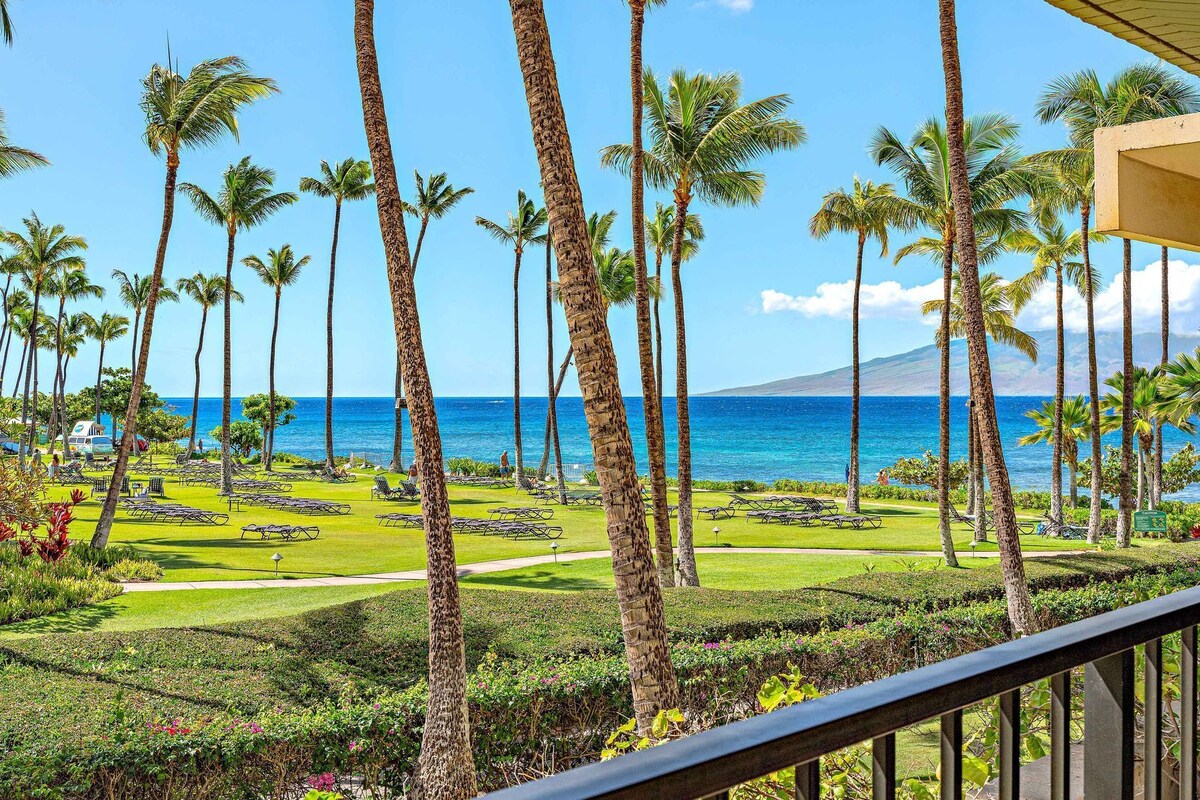 A tropical resort with a view of the ocean, palm trees, and a well-maintained lawn with lounge chairs.