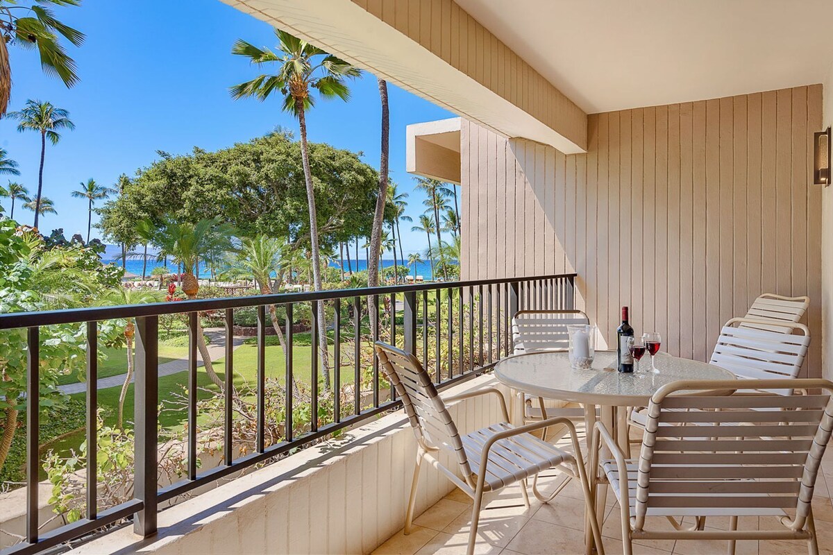 A balcony with a table and chairs, overlooking a tropical landscape with palm trees and a view of the ocean.