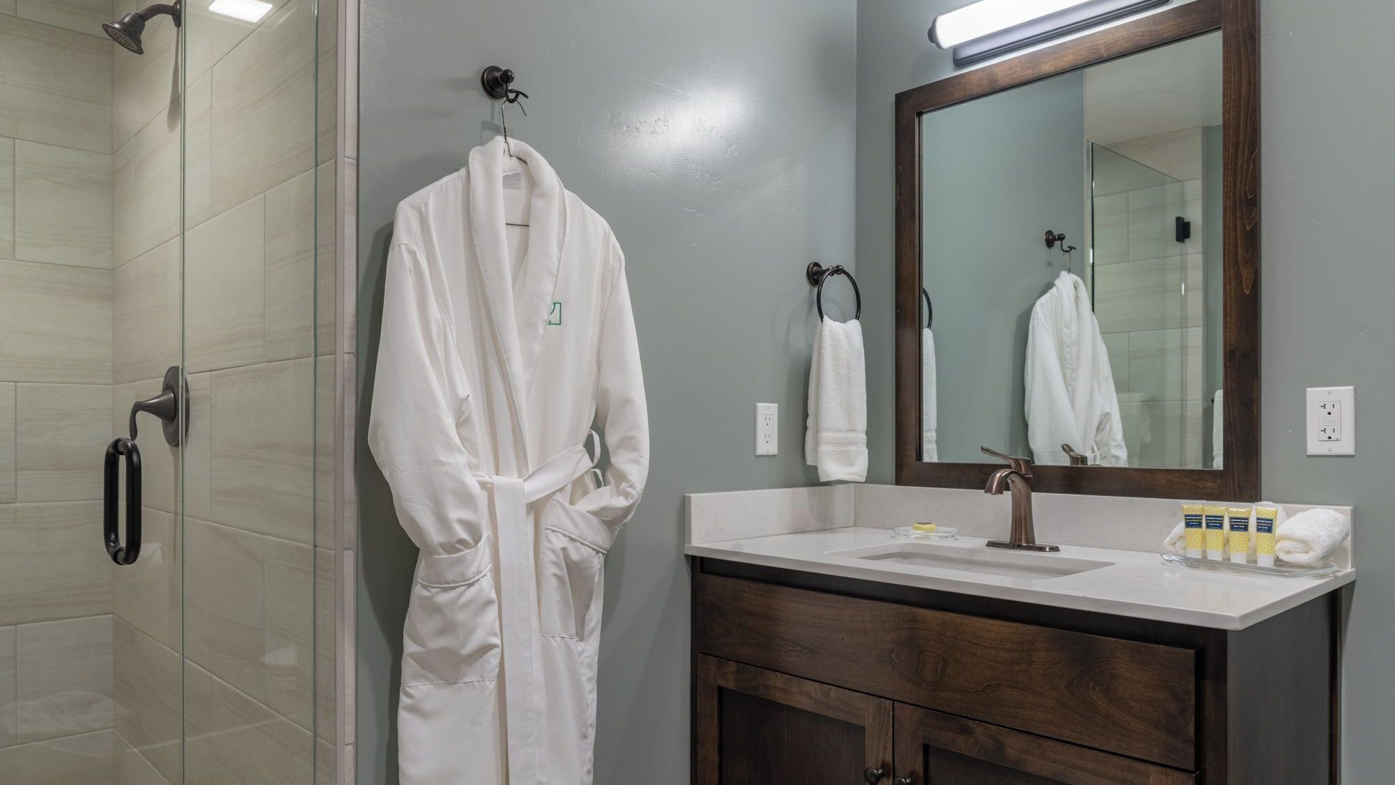 A bathroom with a glass shower enclosure, a white robe hanging on a hook, a wooden vanity with a mirror, and toiletries on the counter.