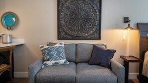 Ein graues Sofa mit gemusterten Kissen, ein Wanddekor, ein kleiner Tisch mit einer Pflanze und eine Lampe.