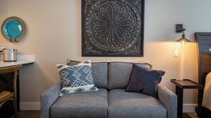A grey sofa with patterned cushions, a decorative wall piece, a small table with a pot, and a lamp. - Paso Park Suites by Avantstay Park-view Balcony Downtown Paso Robles (Paso Robles)