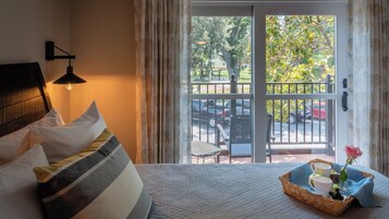 Habitación de hotel con cama, mesita de noche, lámpara, ventana con cortinas y balcón con vista a la calle y automóviles.