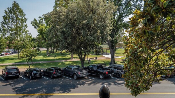 Ein Parkplatz mit mehreren Fahrzeugen, darunter Autos und Lastwagen, sowie ein Grünflächenbereich mit Bäumen im Hintergrund.