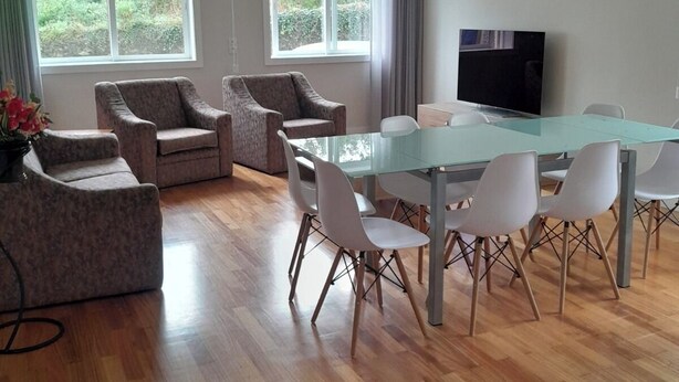 Ein modernes Wohnzimmer mit einem Glastisch, weißen Stühlen, zwei Sofas, einem Flachbildfernseher und großen Fenstern mit Blick auf Grünflächen.