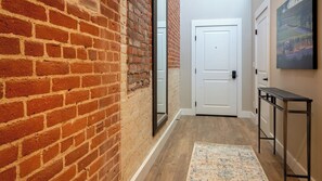 A hallway with a brick wall, a white door, a small table, and a rug. - Paso Park Suites by Avantstay Balcony Over the Square + Walkable Paso Robles (Paso Robles)