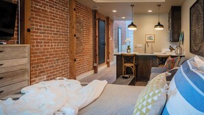 A cozy room with a brick wall, a wooden dresser, a bed with white bedding, a small kitchenette, and a dining area with a bar stool. - Paso Park Suites by Avantstay Balcony Over the Square + Walkable Paso Robles (Paso Robles)