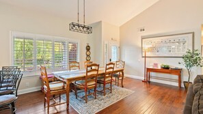 A dining area with a wooden table and chairs, a hanging light fixture, and a view of the outdoors through large windows.