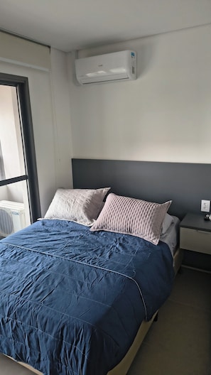 Una habitación de hotel con cama, mesita de noche, equipo de aire acondicionado y ventana.