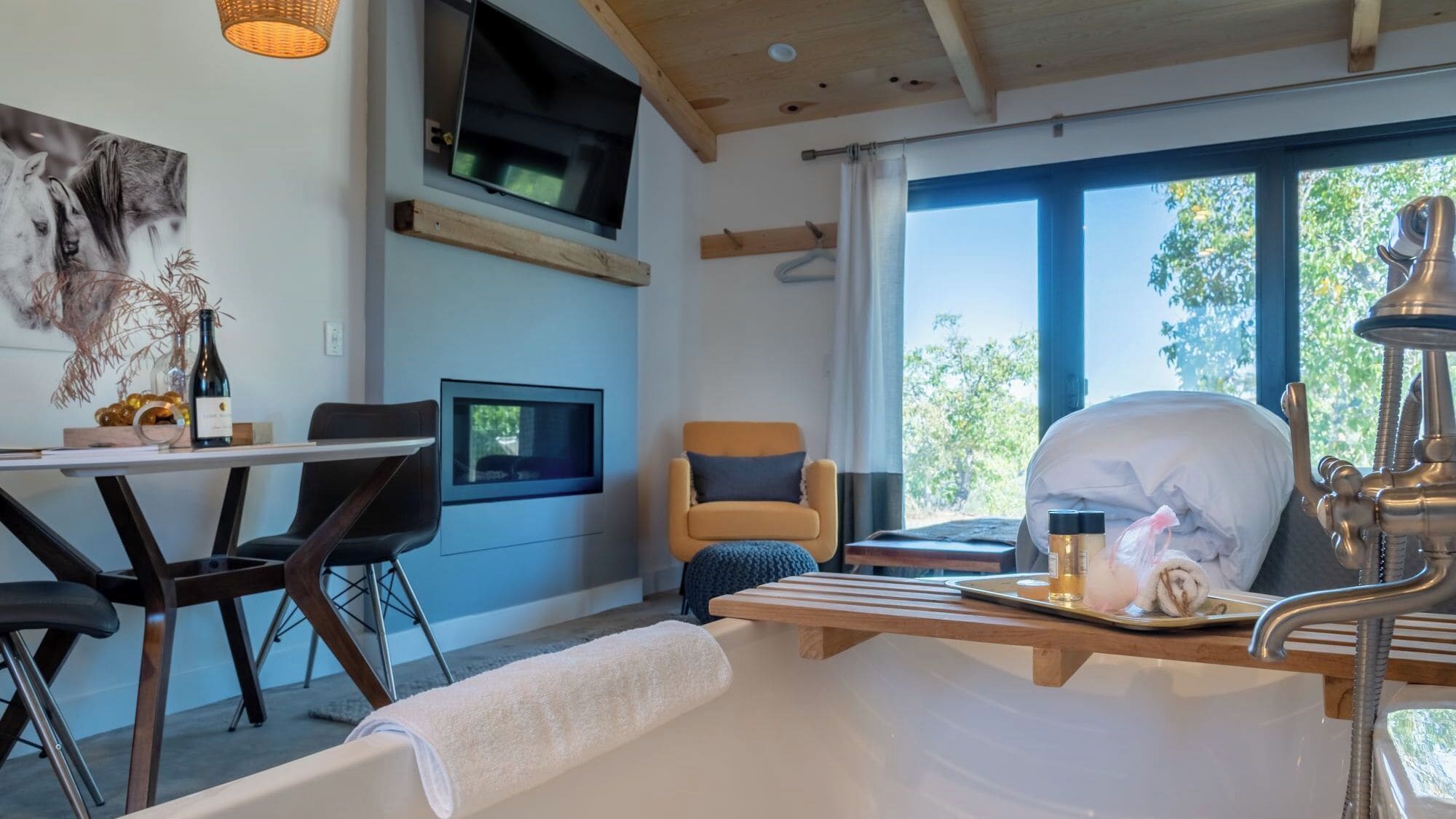 A modern room with a bed, a TV mounted on the wall, a dining area with chairs, and a window with a view of trees.
