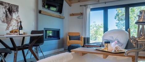 A modern room with a bed, a TV mounted on the wall, a dining area with chairs, and a window with a view of trees.