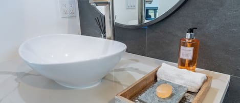 Een badkamer met een wit wastafelbakje, een fles handzeep, een zeepstuk en een handdoek op een houten dienblad.