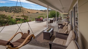 A covered outdoor patio with seating, a swing chair, and a view of the hills. - The J's Nest by Avantstay Hot Tub + BBQ + Lawn Games Panoramic Lake + Oak Hill Views (Bradley)