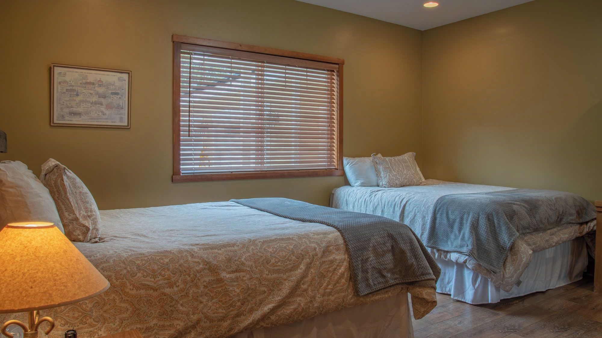 A room with two beds, a window with blinds, a lamp, and a framed picture on the wall.