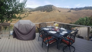 An outdoor dining area with a table set for four, surrounded by chairs with colorful cushions. A grill is covered with a cloth, and a large water dispenser is visible. The setting overlooks a hilly landscape. - The J's Nest Cottage by Avantstay Canyon Views + BBQ Paso Robles Wine Country (Paso Robles)