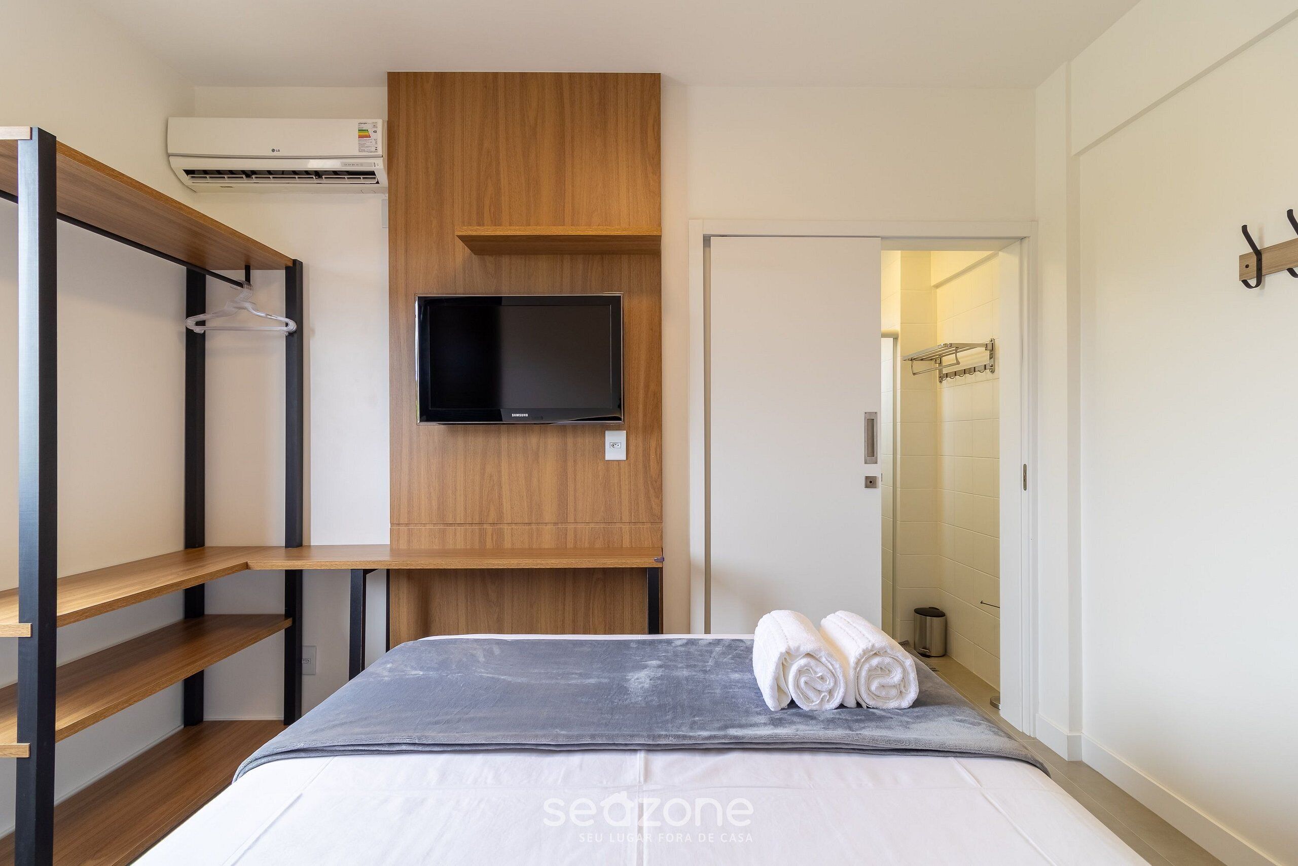 A modern bedroom with a bed, a TV mounted on a wooden panel, and a wardrobe with a hanging rack.