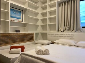 Ein Hotelzimmer mit einem Bett, einem Holz-Kopfteil, einem Regal mit einem gerahmten Bild, einem Fenster mit Vorhängen und einem Zitat an der Wand.