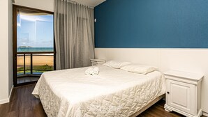 A bedroom with a large bed, a nightstand, and a view of the beach through a window. - Apto With Panoramic View of the sea Front Por204 (Florianópolis)
