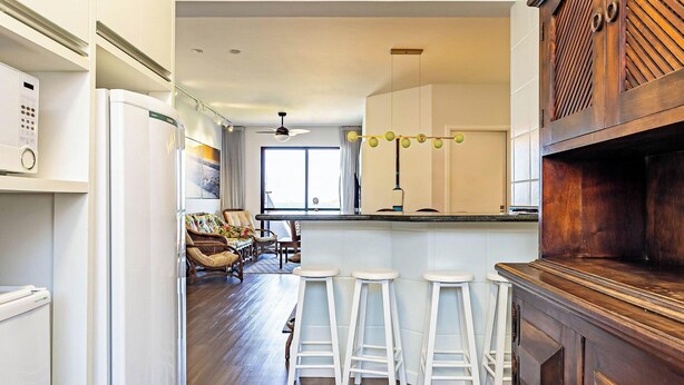 A modern kitchen with white cabinetry, a wooden bar area, and a seating area with a view of the outdoors. - Apto With Panoramic View of the sea Front Por204 (Florianópolis)