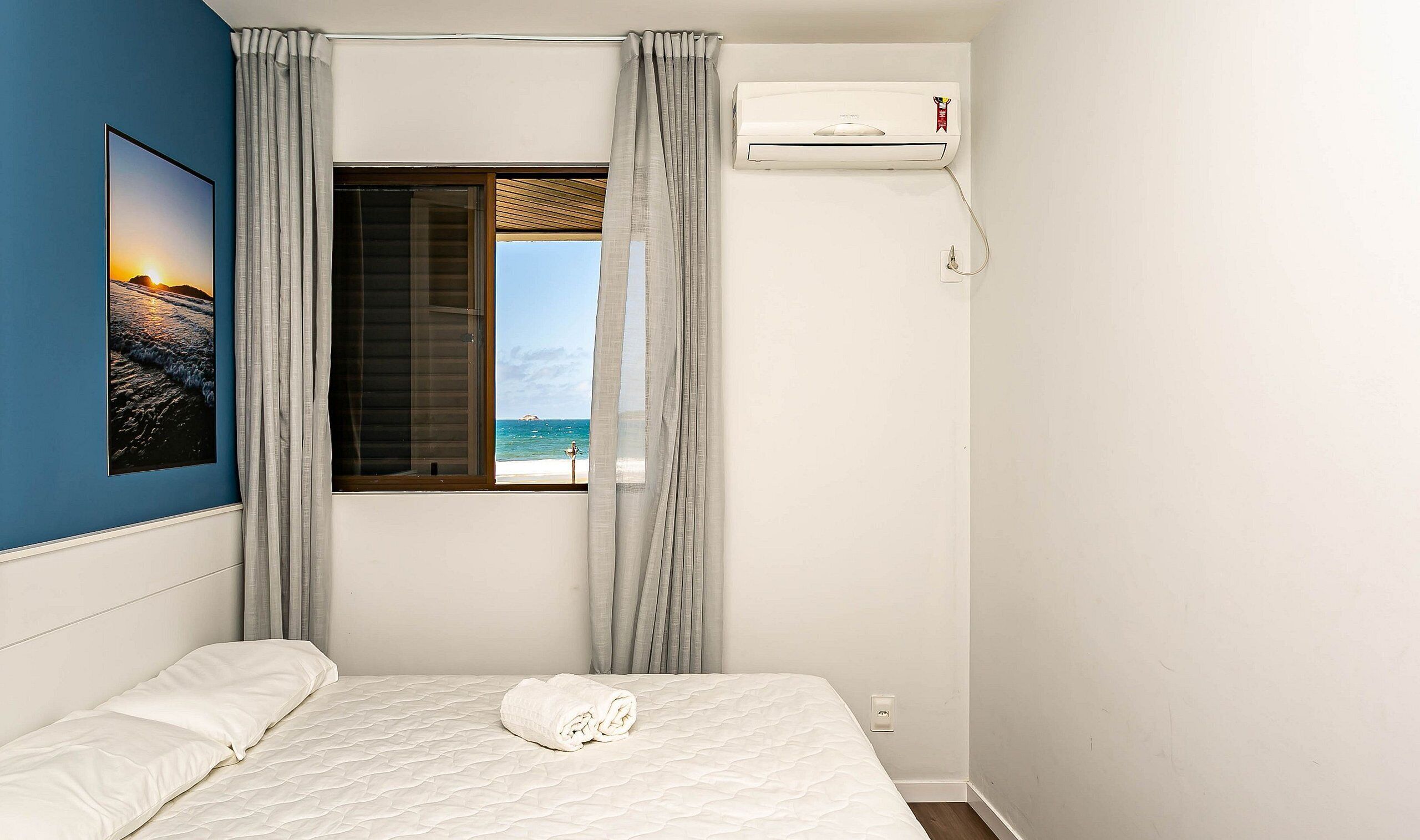 A bedroom with a bed, a window with a view of the sea, an air conditioning unit, and a painting on the wall.