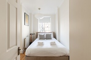 Ein schmales Hotelzimmer mit einem Bett, einem Heizkörper, einer Stehlampe und einem Fenster mit Blick auf Gebäude.
