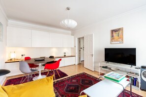 Ein modernes Wohnzimmer mit Flachbildfernseher, eine Essecke mit Tisch und Stühlen sowie eine Küchenzeile mit weißen Schränken.